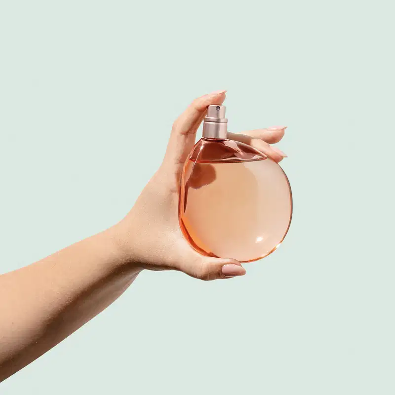 Mão feminina segurando um frasco arredondado em tons de rosa em fundo azul claro.
