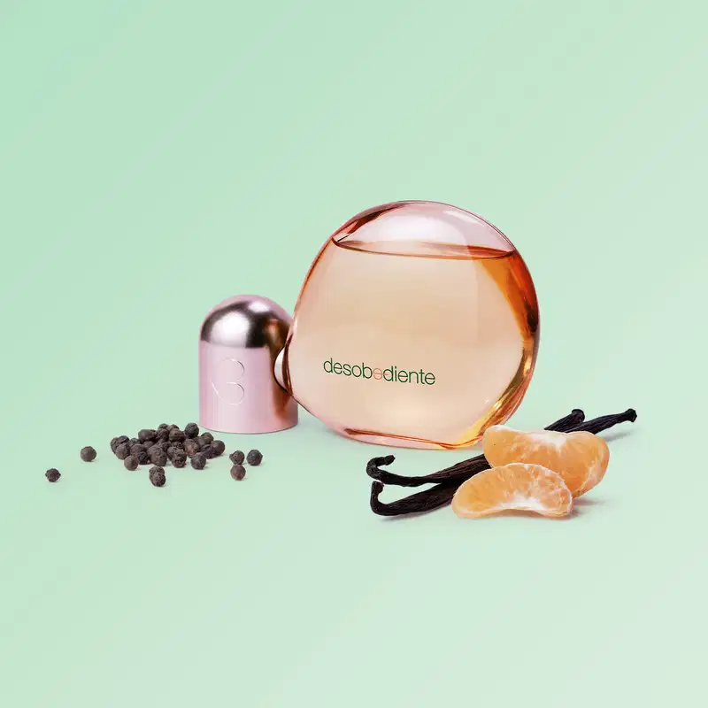 Frasco de Eau de Parfum Desobediente Quem Disse, Berenice? com design arredondado em tom de rosa, sobre superfície verde clara, acompanhado de fatias de laranja e vagens de baunilha.