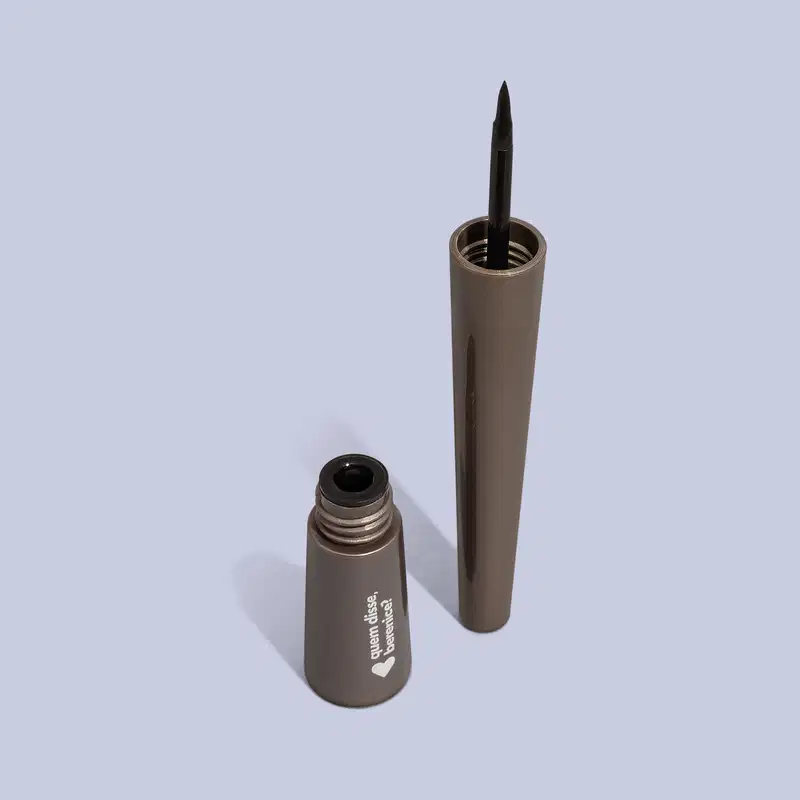Produto de beleza em embalagem metálica cinza escuro, um frasco e um lápis. O frasco possui um rótulo com um coração, uma ponta preta e uma extremidade oca.