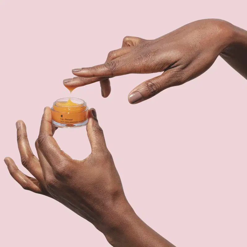Mãos femininas, uma segurando um frasco aberto de balm labial de hidratação intensa Lip, Please! Quem Disse, Berenice? e a outra com uma pequena quantidade do produto em um dos dedos em fundo lilás claro.