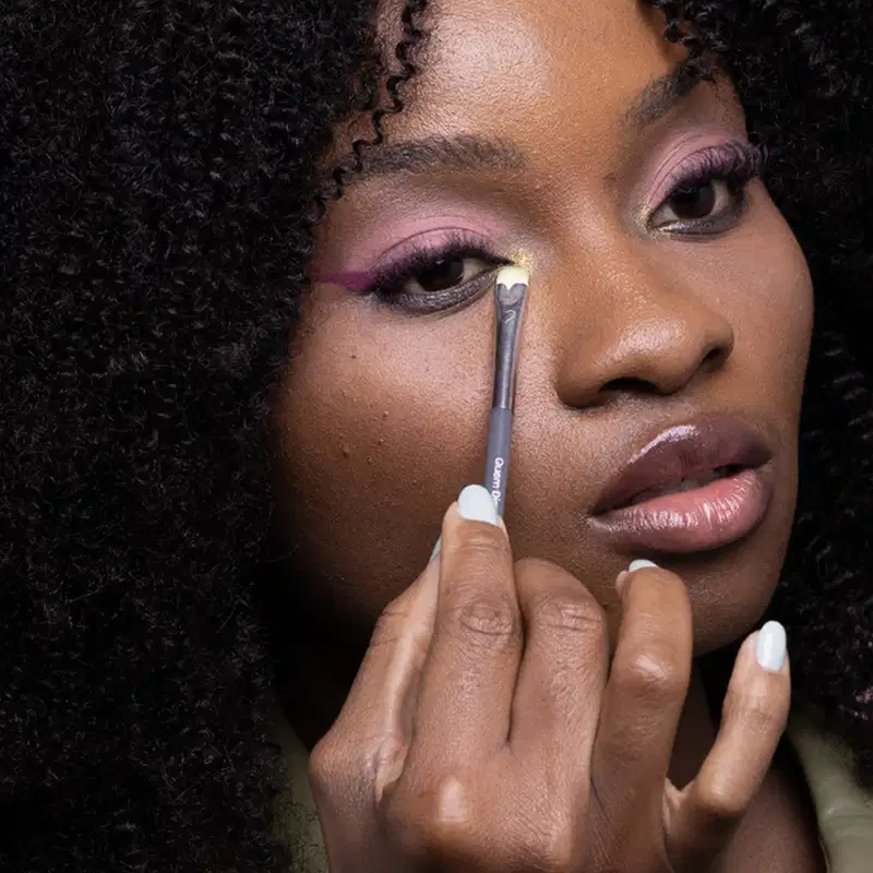 Mulher de pele negra aplicando sombra rosa em seu rosto, com cabelos pretos e unhas esmaltadas em branco.