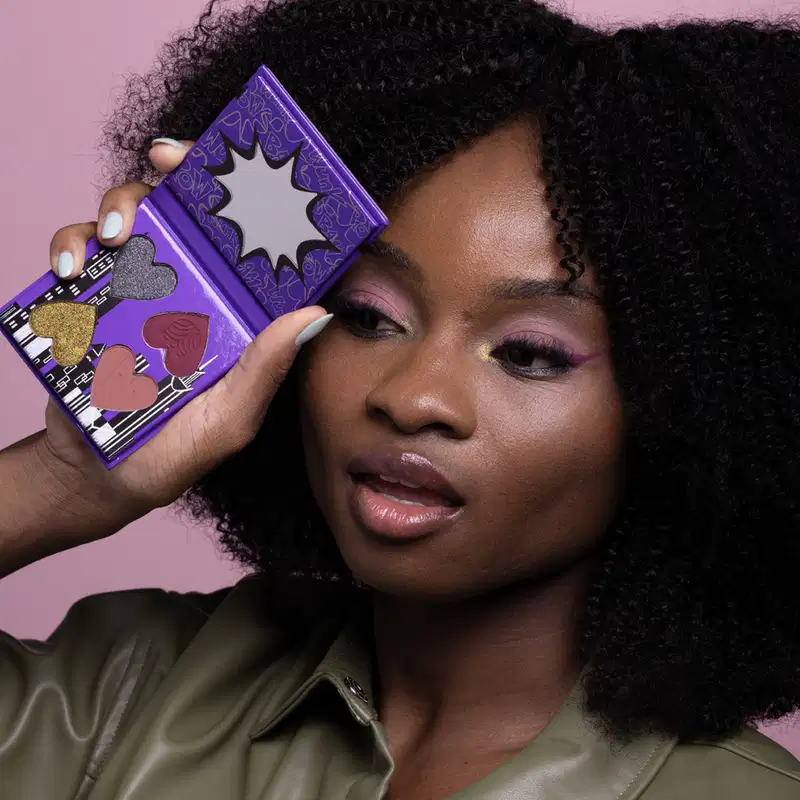 Uma mulher de pele negra segura uma mini paleta de maquiagem da marca Meninas Superpoderosas Quem Disse, Berenice? tons de azul e rosa, em um fundo rosa.