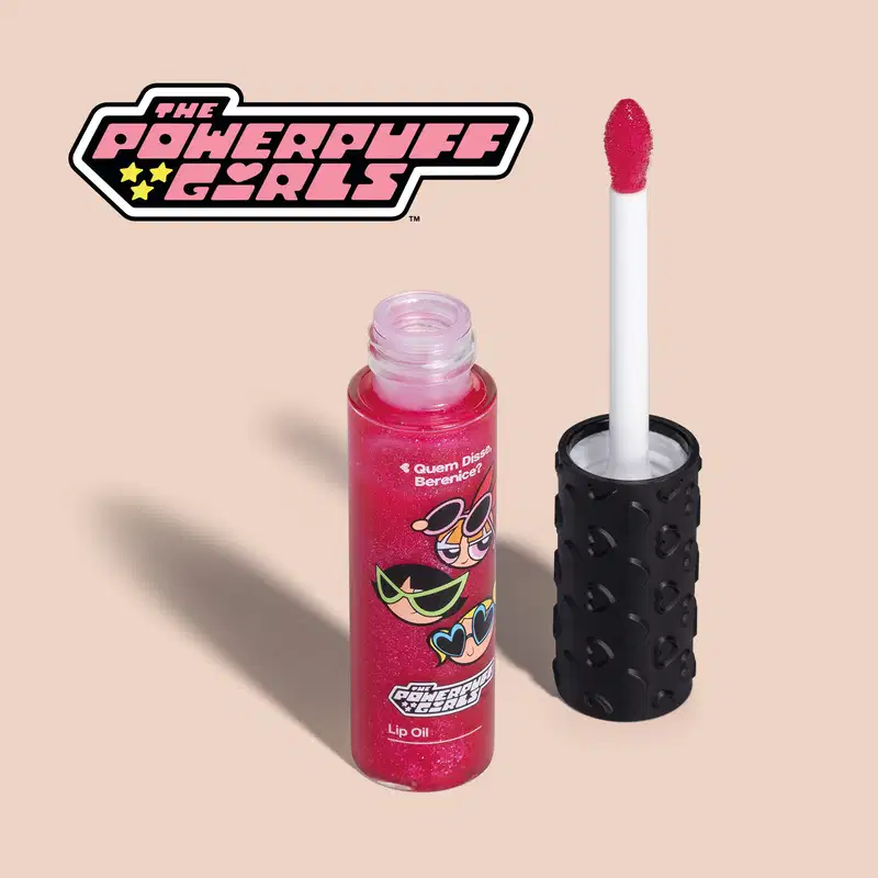 Frasco de lip oil na cor rosa da marca Meninas Superpoderosas Quem Disse, Berenice? em embalagem transparente mostrando a tonalidade do produto, tampa preta com haste e esponjinha vermelha, mostrando ilustração de personagens, em fundo rosa.