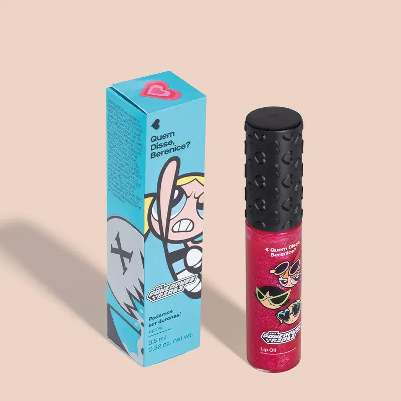 Embalagem azul com ilustração de personagem das Meninas Superpoderosas, contendo um lip oil rosa em embalagem transparente e tampa preta em ambiente liso e claro.