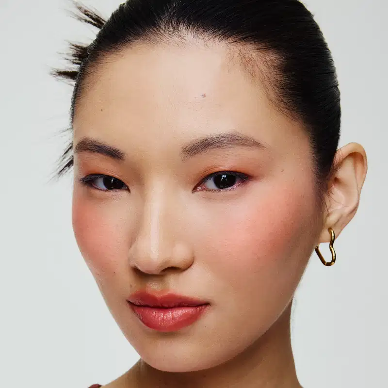 Rosto de uma mulher asiática com cabelo escuro e penteado elegante. Ela usa brincos dourados e maquiagem com tons de laranja e vermelho nos olhos e lábios. O fundo é branco, criando um ambiente minimalista e focado no rosto da modelo.