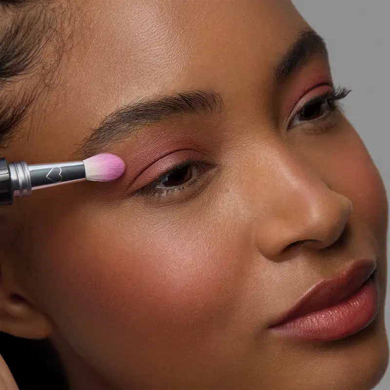 Uma pessoa aplica um produto de maquiagem rosa em sua pálpebra com um pincel. O rosto da pessoa está em primeiro plano, com pele morena e lábios rosados. O fundo é neutro, permitindo o destaque do produto de beleza em uso.