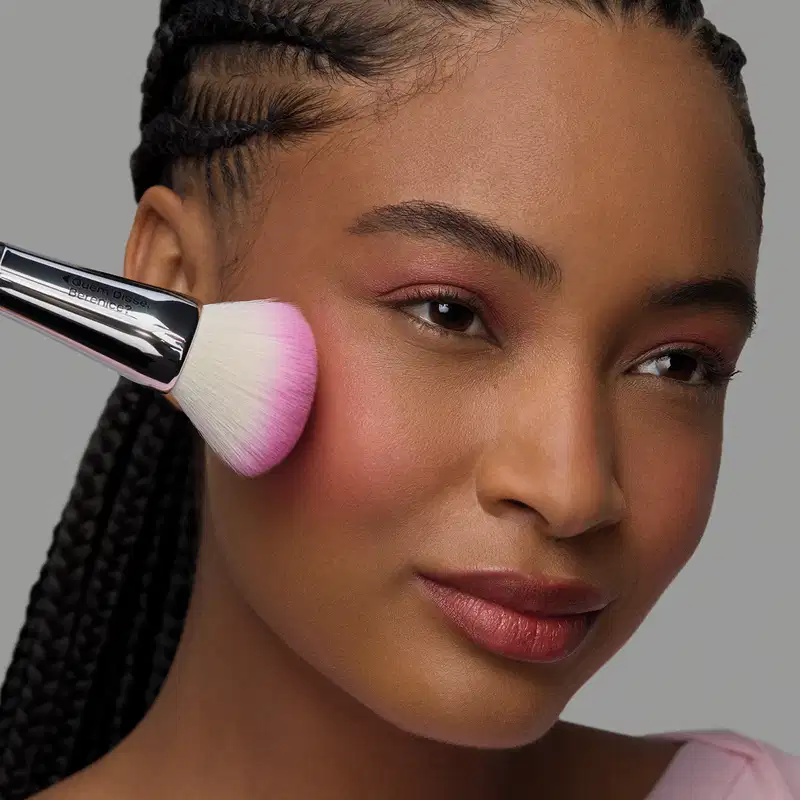 Uma pessoa com penteado trançado aplica um item de perfumaria rosa em seu rosto. Ela tem expressão séria e olhar direto. O fundo é cinza, criando um ambiente minimalista e focado no produto.