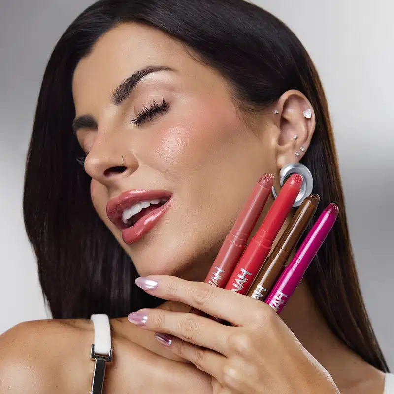 A mulher segura vários produtos de maquiagem da marca, incluindo batons em tons de vermelho e rosa, em um ambiente de fundo cinza, sugerindo um contexto de aplicação e uso de produtos de beleza.