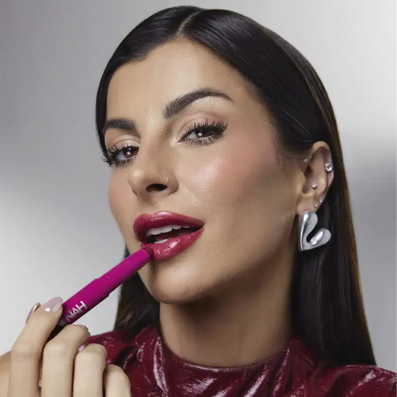 A mulher aplica um batom rosa em seus lábios, com maquiagem elaborada e acessórios de joias, em um ambiente minimalista e monocromático, sugerindo um contexto de produto de beleza da marca Nah.