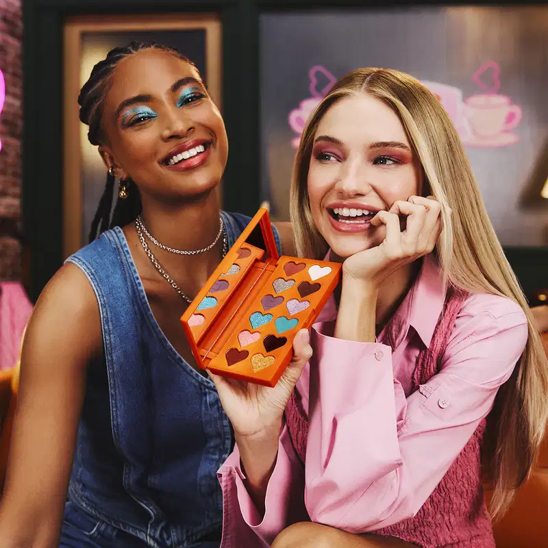 Duas mulheres sorrindo em ambiente decorado com elementos rosa e roxo, sugerindo um cenário relacionado a cafeteria. Uma mulher segura uma paleta laranja com corações coloridos, usando roupas em tons de rosa. O ambiente parece acolhedor e alegre.