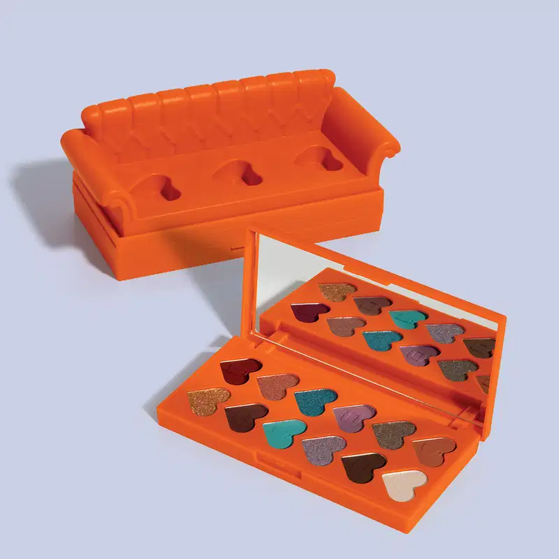 Paleta laranja em forma de sofá com compartimentos em formato de coração, contendo sombras de olho em tons de marrom, dourado e turquesa. A caixa possui uma tampa com encaixes. 