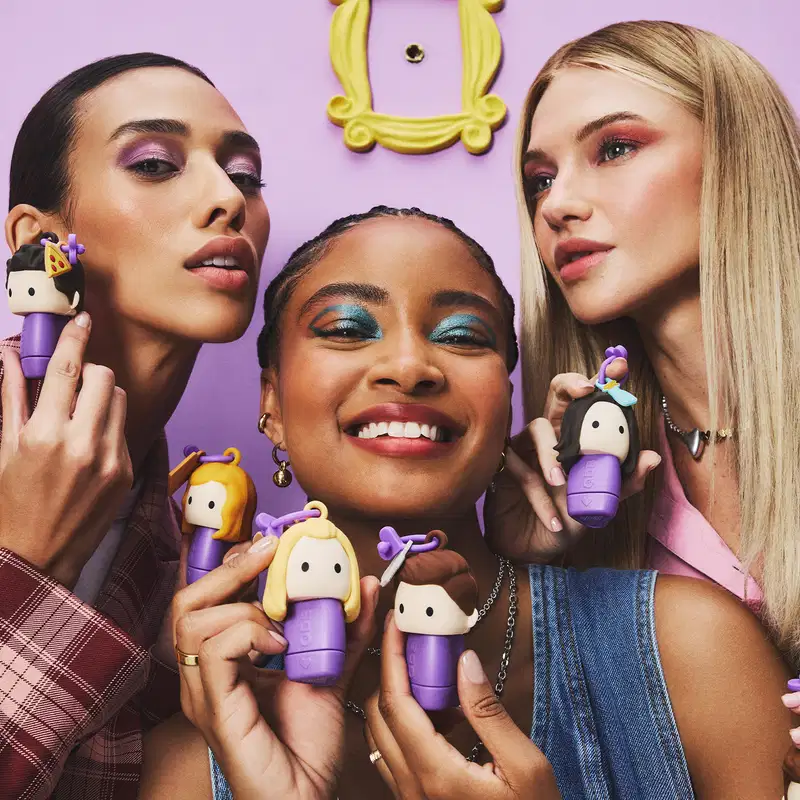 Três mulheres segurando os batons em um ambiente roxo e amarelo. Elas usam maquiagem colorida e acessórios combinando com os itens. O cenário sugere um contexto de beleza e cuidados pessoais.