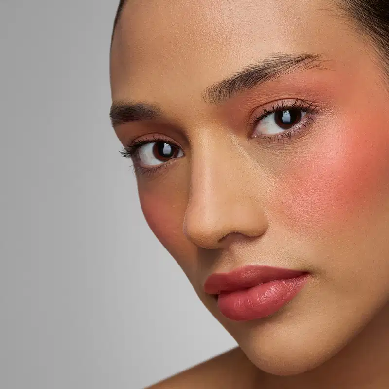 Rosto de uma pessoa com pele negra, olhos escuros e expressão séria. Lábios vermelhos e sobrancelhas bem definidas. Fundo cinza neutro que destaca os traços faciais. Imagem de alta qualidade que sugere um ambiente de beleza ou cosmética.