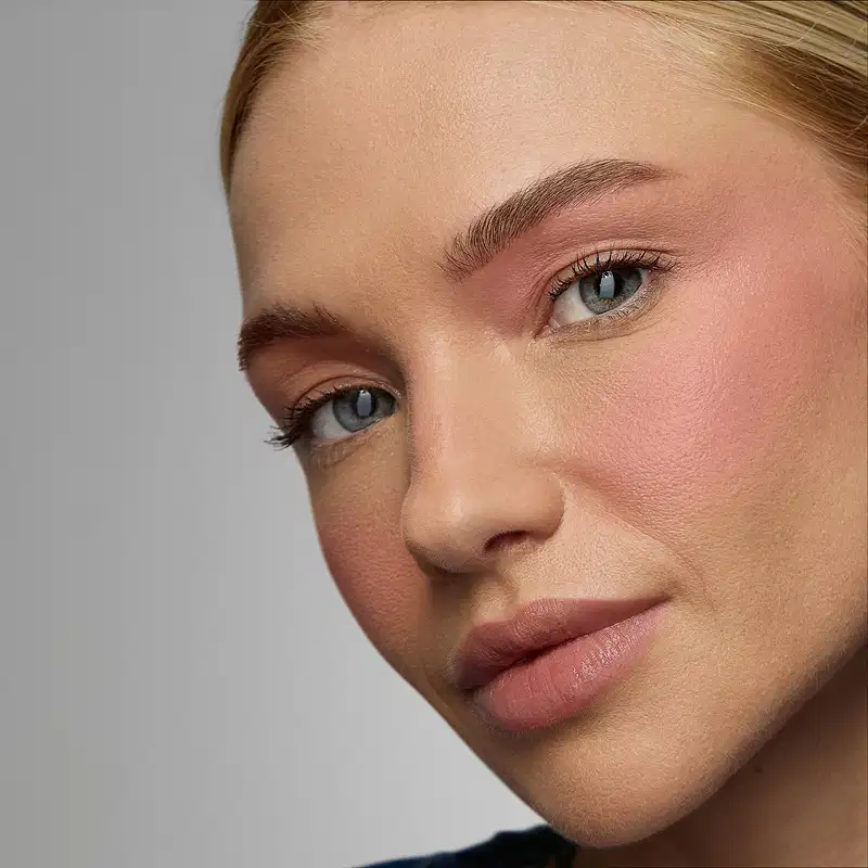 Rosto de uma mulher com olhos azuis, sobrancelhas bem definidas e lábios rosados. A iluminação suave e o fundo neutro criam um ambiente elegante e sofisticado, típico de uma campanha de produtos de beleza.