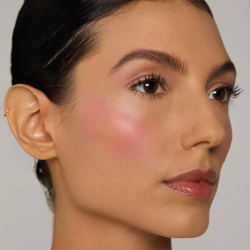 Rosto feminino com maquiagem natural, cabelos escuros e pele corada, em ambiente com fundo branco e iluminação suave.