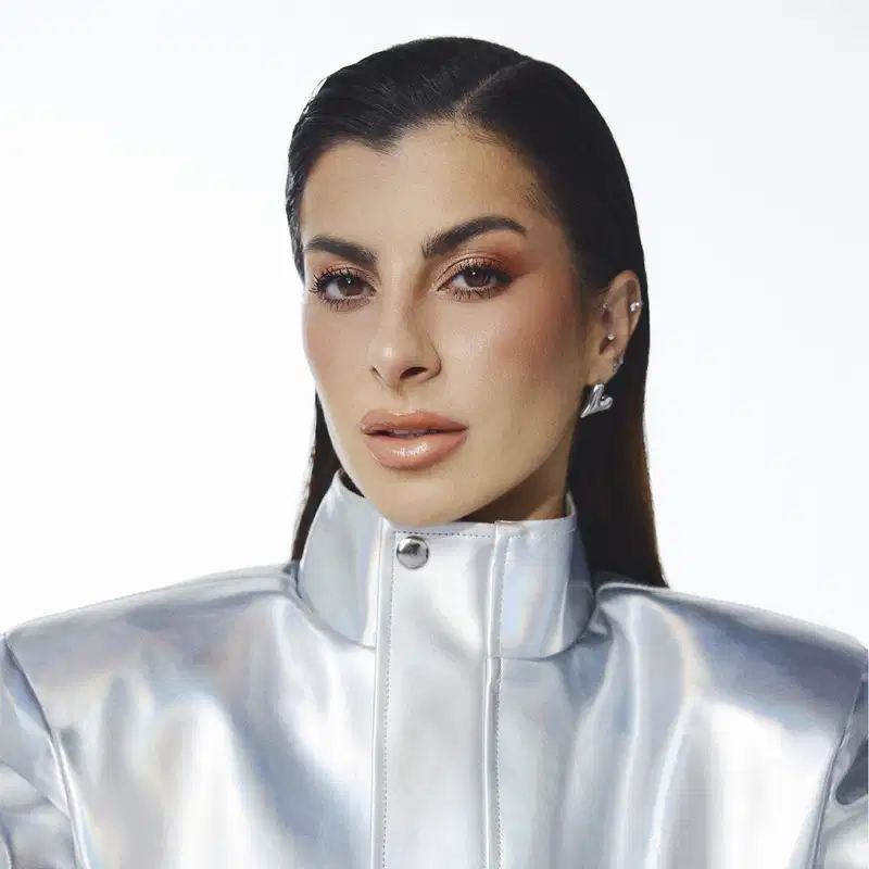 
Mulher com cabelos escuros e maquiagem em close-up, usando jaqueta prateada, em fundo branco.
