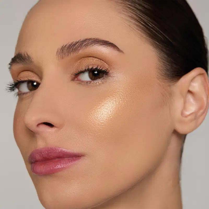 Rosto de mulher com maquiagem, cabelo escuro penteado para trás, fundo cinza claro, iluminação suave.
