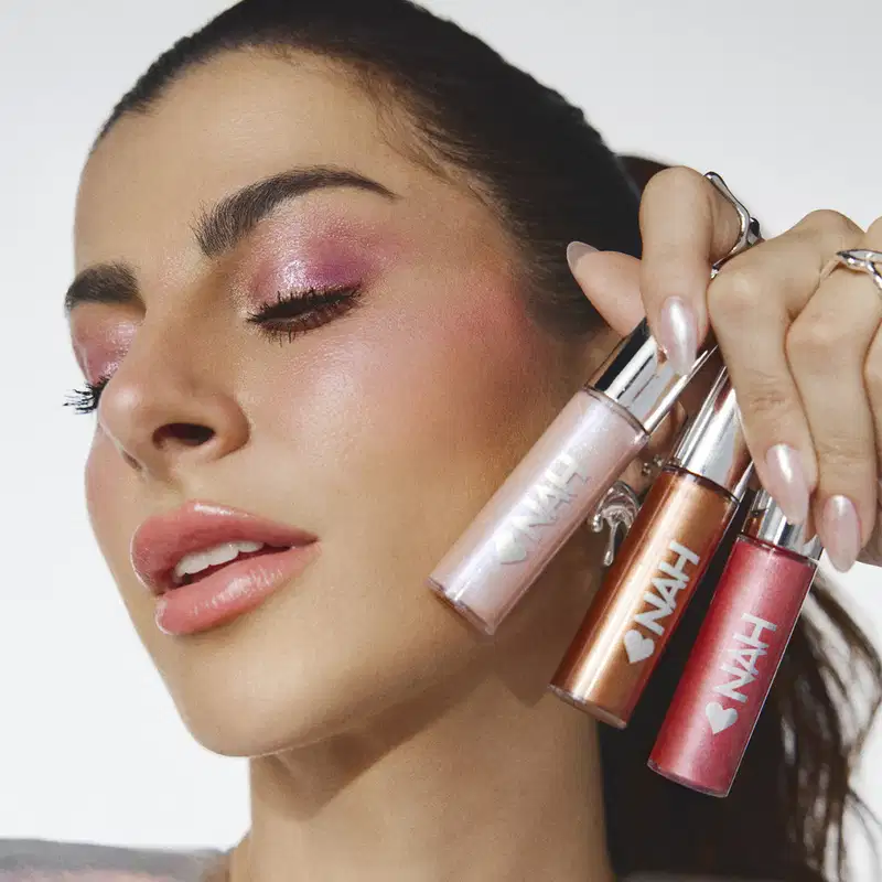 Imagem de close-up de uma mulher segurando três itens de make da marca Nah Cardoso, com embalagens em tons de rosa, em um fundo branco.