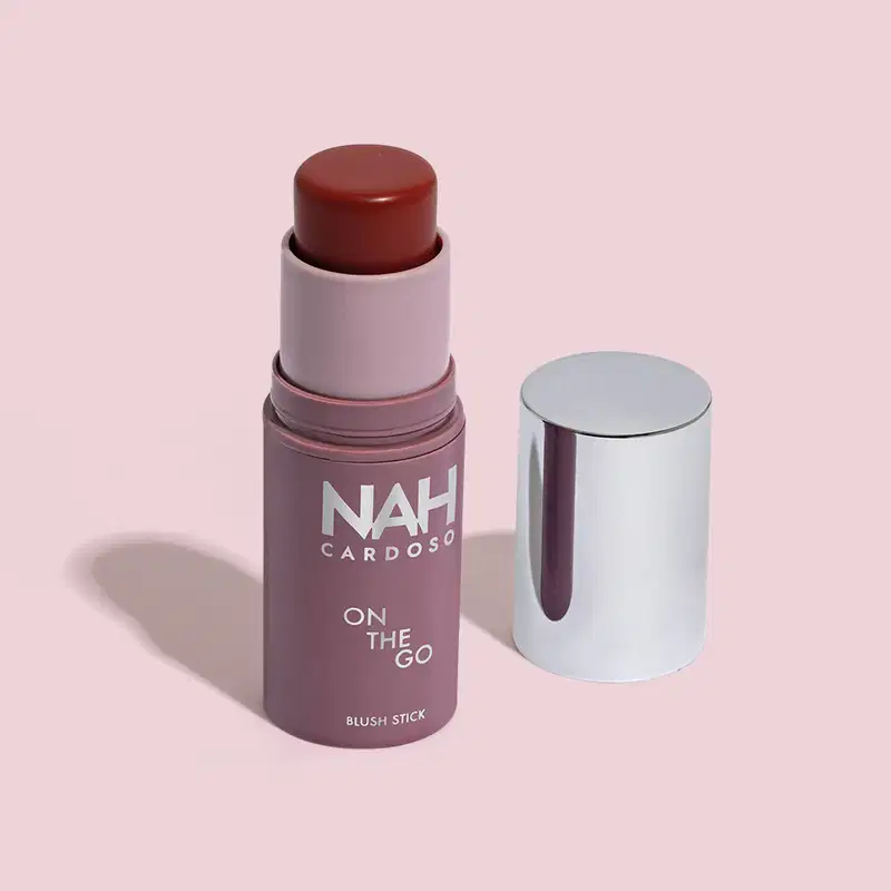 Produto de beleza em formato de bastão, com embalagem roxa e branca, exibindo o nome "NAH CARDOSO" e o texto "ON THE GO BLUSH STICK". O produto possui uma tampa prateada e uma cor marrom escuro.