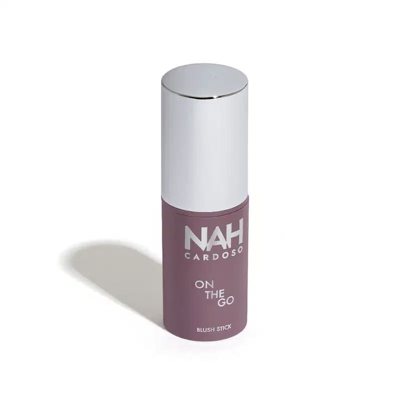 Produto de beleza em embalagem cilíndrica branca e roxa, com o nome "NAH CARDOSO" e a descrição "ON THE GO BLUSH STICK" impresso no rótulo. Trata-se de um blush em bastão, com design compacto e portátil, ideal para uso rápido e prático.