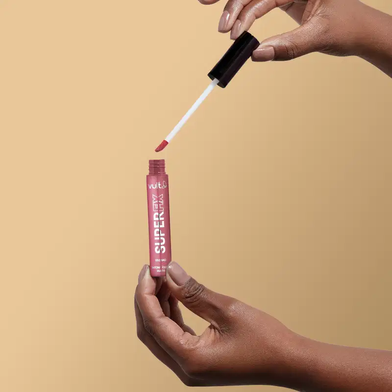 A imagem mostra uma mão segurando o produto aberto do Batom Líquido da marca Vult, na cor rosa queimado. A embalagem do produto é um tubo vermelho com o nome da marca "Vult" impresso.