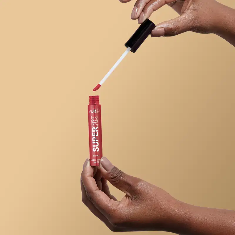A imagem mostra uma mão segurando o produto aberto do Batom Líquido da marca Vult, na cor vermelha. A embalagem do produto é um tubo vermelho com o nome da marca "Vult" impresso.