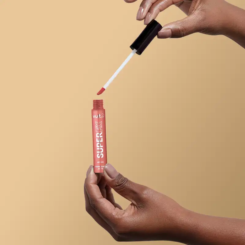 A imagem mostra uma mão segurando o produto aberto do Batom Líquido da marca Vult, na cor castanho. A embalagem do produto é um tubo vermelho com o nome da marca "Vult" impresso.