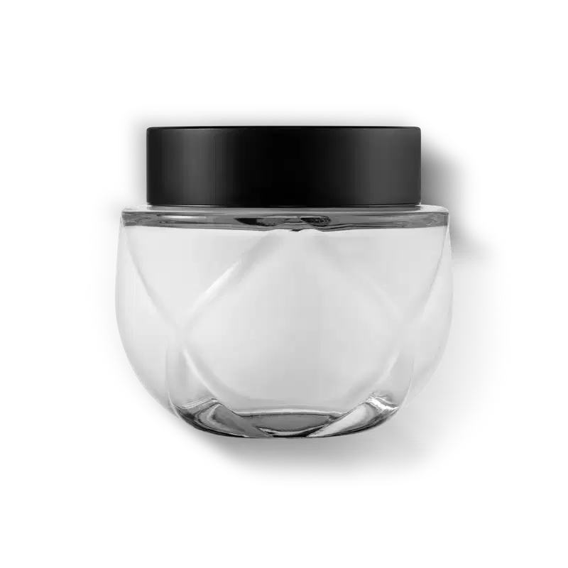 Frasco de vidro transparente arredondado com tampa preta. A embalagem tem um design minimalista e clean, com linhas suaves e uma forma esférica. As cores predominantes são o preto da tampa e o transparente do vidro e a cor branca do creme.