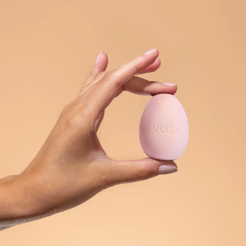 Mãos segurando uma esponja de maquiagem Vult em um fundo laranja, sugerindo um ambiente de cuidados com a pele e produtos de beleza.