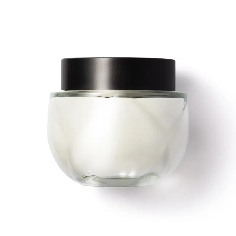 Frasco de vidro arredondado, transparente com tampa preta de rosquear, contendo o creme na cor branca. Design minimalista e elegante.