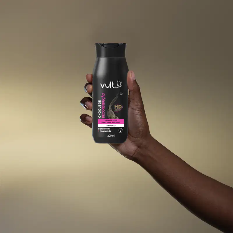 A imagem apresentada é uma mão segurando o produto, o shampoo da marca Vult. A embalagem é um tubo preto com o logotipo da marca e informações sobre o produto, como "Choque de Reconstrução" e "Tecnologia HD".