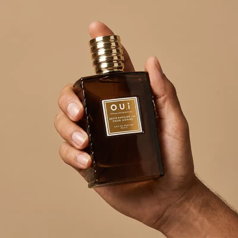 Recipiente de item de perfumaria da marca O.U.i em mãos masculinas sobre fundo bege, com detalhes dourados na embalagem.