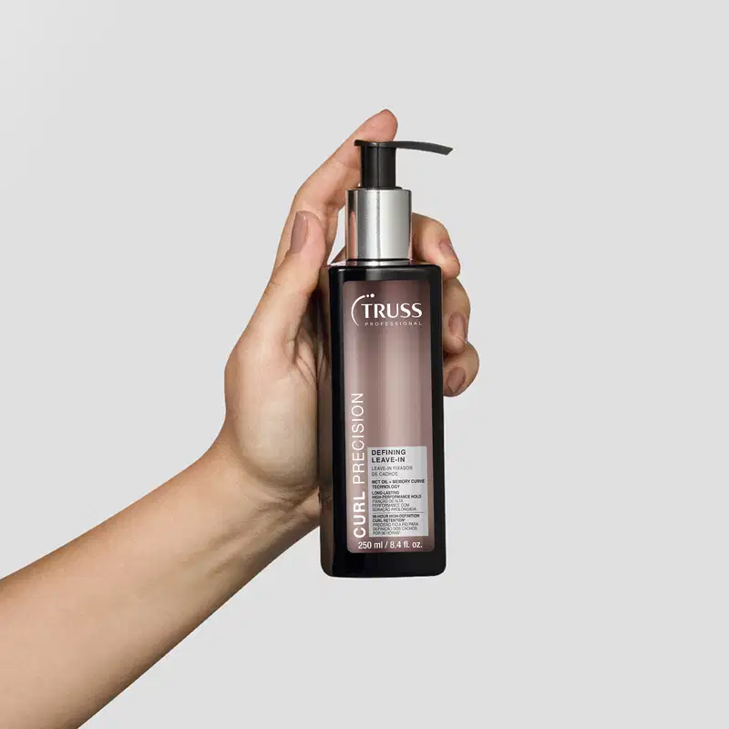 Frasco preto de item de cabelos da marca Truss com bomba doseadora, rótulo com nome "Curl Precision" e texto indicando que é um leave-in finalizador, sendo segurado por uma mão feminina.