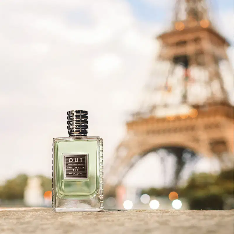 Frasco de item de perfumaria da marca O.U.i em primeiro plano, com a Torre Eiffel desfocada ao fundo, em um cenário urbano com luzes de fundo.