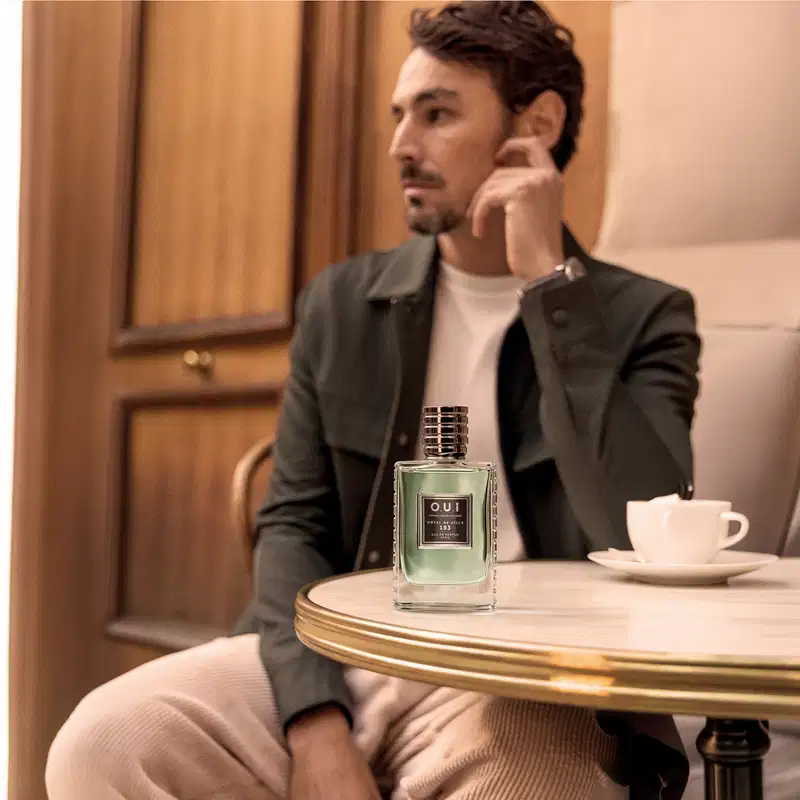 Um homem de terno cinza está sentado em uma mesa de madeira, com uma xícara de café e um frasco de perfumaria da marca O.U.i em primeiro plano.
