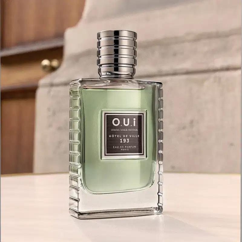 Frasco de item de perfumaria da marca O.U.i em embalagem de vidro transparente com detalhes em prateado, posicionado sobre superfície clara em ambiente com paredes de pedra.
