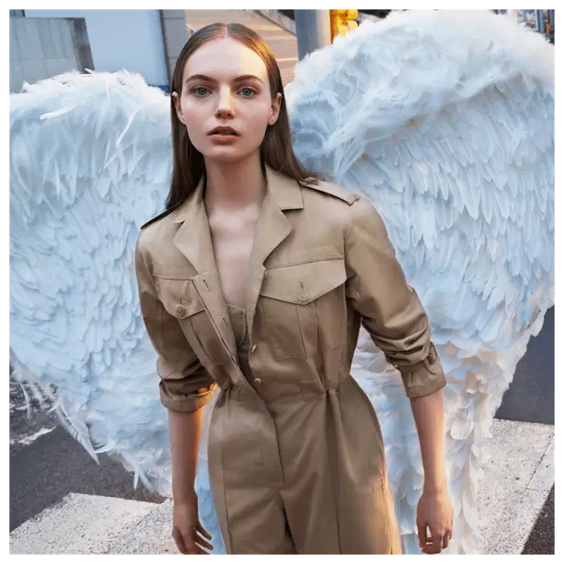 Ambiente urbano com asas de anjo brancas ao fundo, modelo vestindo trench coat bege da marca Burberry, em uma composição que sugere uma campanha de moda da marca.