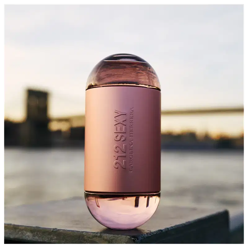 Frasco de perfume Carolina Herrera 212 Sexy, com formato cilíndrico e tampa arredondada, em tons de rosa claro. O rótulo exibe o nome da fragrância em letras prateadas.
