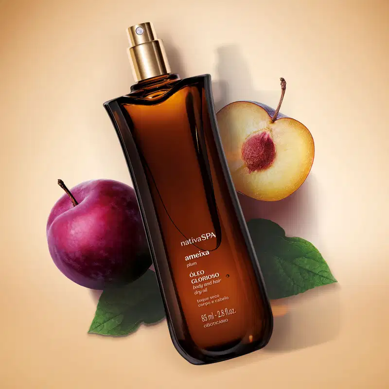 Frasco alongado na cor vinho escuro com borrifador dourado e letras brancas de óleo multifuncional glorioso corpo e cabelos Nativa SPA Ameixa Plum sobre fundo bege, acompanhado de ameixa roxa inteira, ameixa cortada ao meio e folhas verdes.