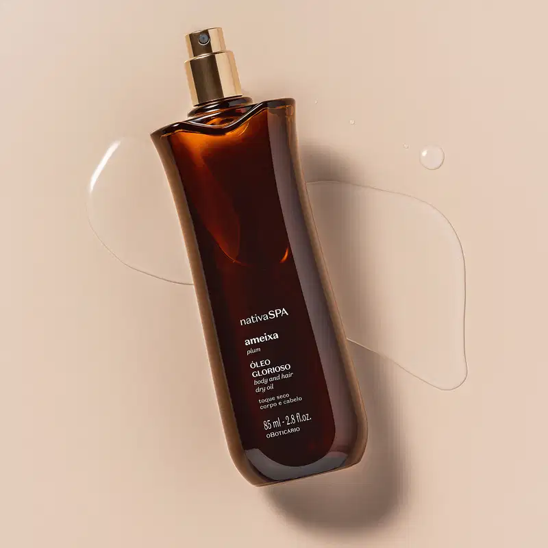 Frasco alongado na cor vinho escuro com borrifador dourado e letras brancas de óleo multifuncional glorioso corpo e cabelos Nativa SPA Ameixa Plum posicionado em superfície bege com gotas de líquido ao redor e sombra projetada.