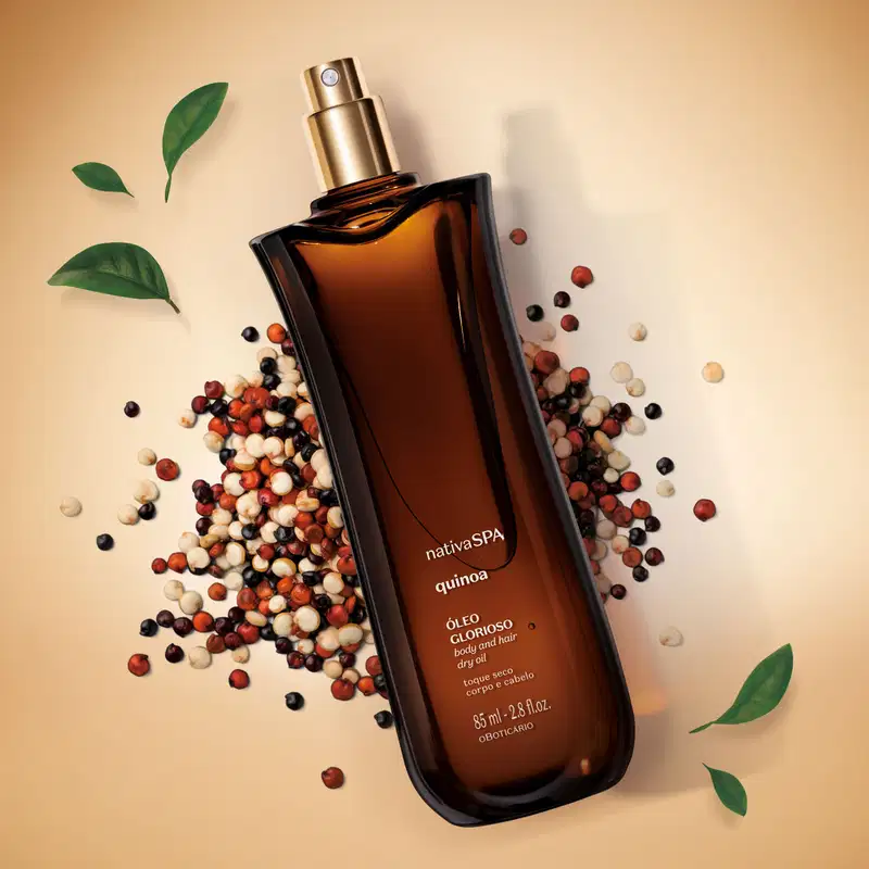 Vidro marrom com pulverizador dourado e letras brancas contendo óleo multifuncional glorioso corpo e cabelos da marca Nativa SPA Quinoa sobre fundo bege, cercado por contas coloridas em tons de vermelho, branco e preto, e folhas verdes decorativas.