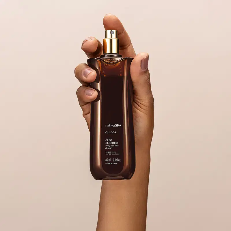 mão segurando um vidro alongado marrom com pulverizador dourado e letras brancas contendo óleo multifuncional glorioso corpo e cabelos da marca Nativa SPA Quinoa em fundo bege.