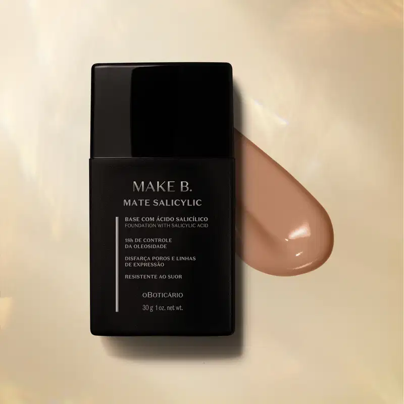 Frasco preto com letras cinzas de base líquida na cor 180 da marca Make B. Mate Salicylic sobre superfície bege com gota de produto bege escuro ao lado, mostrando textura e tonalidade.