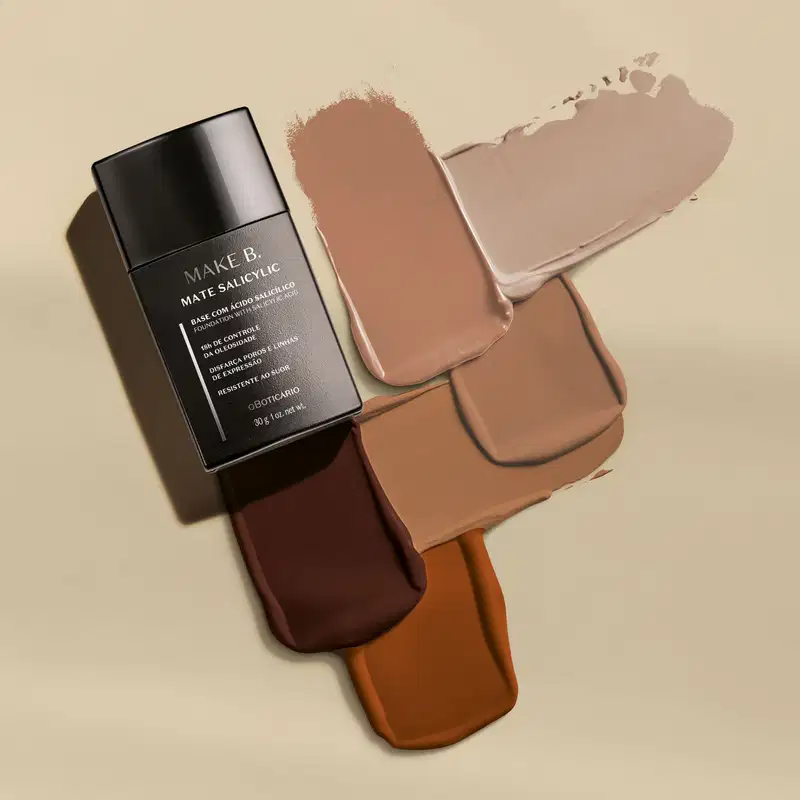 Frasco preto de base líquida Make B. Mate Salicylic ao lado de swatches de todos os tons do produto sobre superfície bege claro.