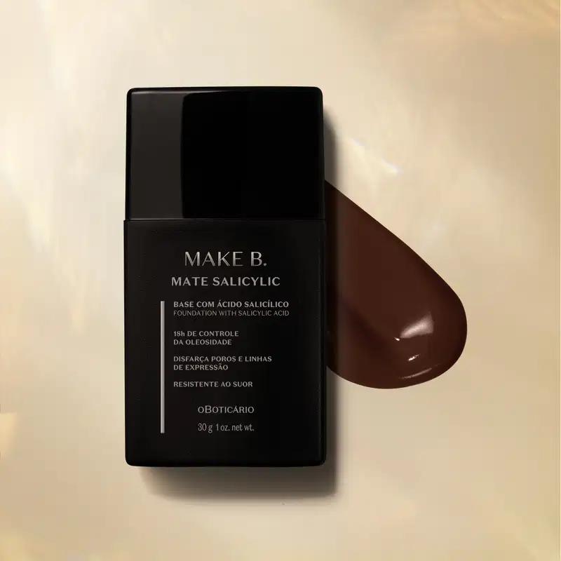 Frasco preto com letras cinzas de base líquida na cor 330 da marca Make B. Mate Salicylic sobre superfície bege com gota de produto marrom escuro ao lado, mostrando textura e tonalidade.