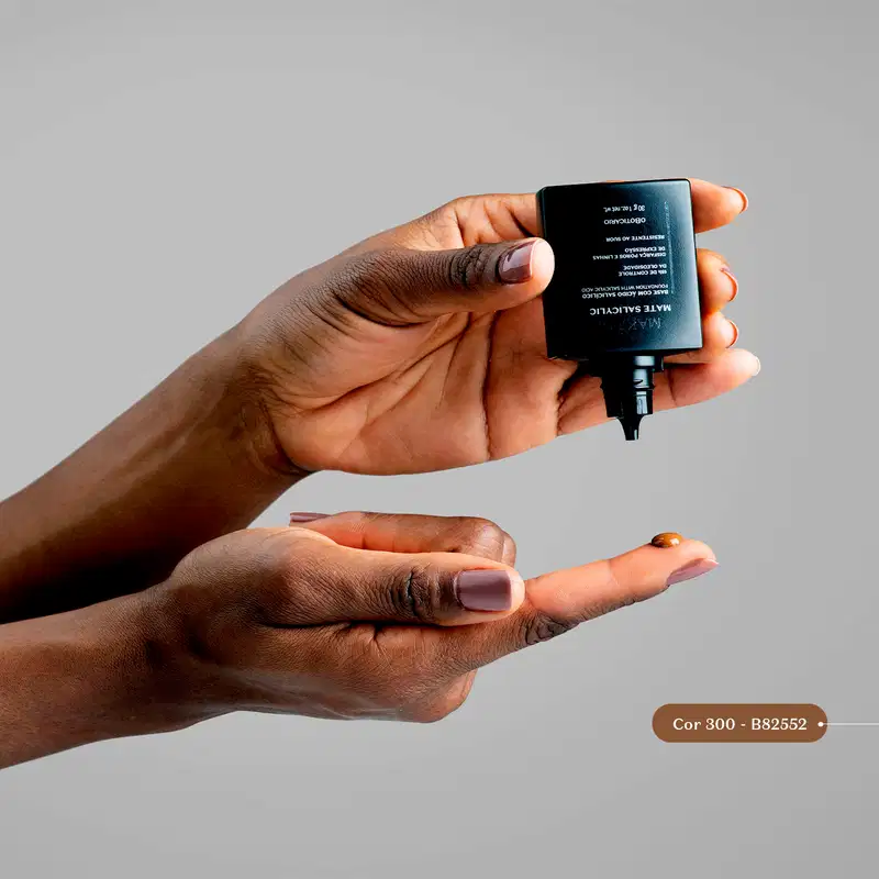 Mão segurando frasco preto da base líquida na cor 330 da marca Make B. Mate Salicylic e aplicando uma gota do produto no dedo da oura mão, sobre fundo azul acinzentado.