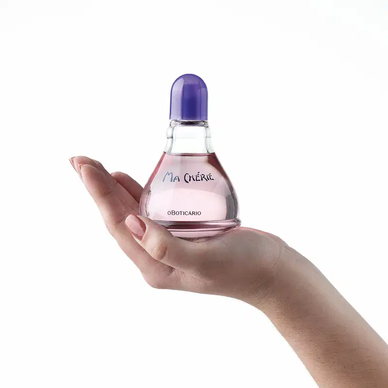 Mão feminina segurando um vidro transparente com fundo arredondado, líquido rosa, tampa e letras azuis, contendo 100ml de desodorante colônia Ma Chérie.