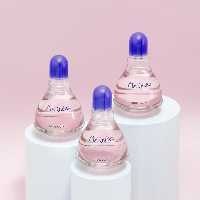 Três vidros transparentes com fundos arredondados, líquidos rosas, tampas e letras azuis de desodorante colônia Ma Chérie. dispostos em pedestais brancos contra fundo rosa claro.