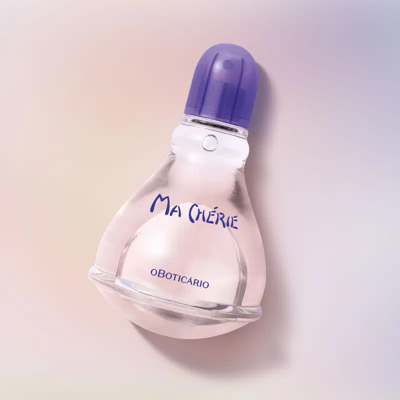 Vidro transparente com fundo arredondado, líquido rosa, tampa e letras azuis, contendo 100ml de desodorante colônia Ma Chérie posicionado sobre superfície neutra em tons pastel rosa e lilás, com sombra suave ao fundo.