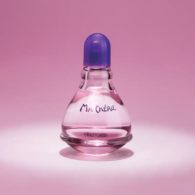 Vidro transparente com fundo arredondado, líquido rosa, tampa e letras azuis, contendo 100ml de desodorante colônia Ma Chérie, posicionado em superfície rosa com sombra refletida, fundo rosa uniforme.